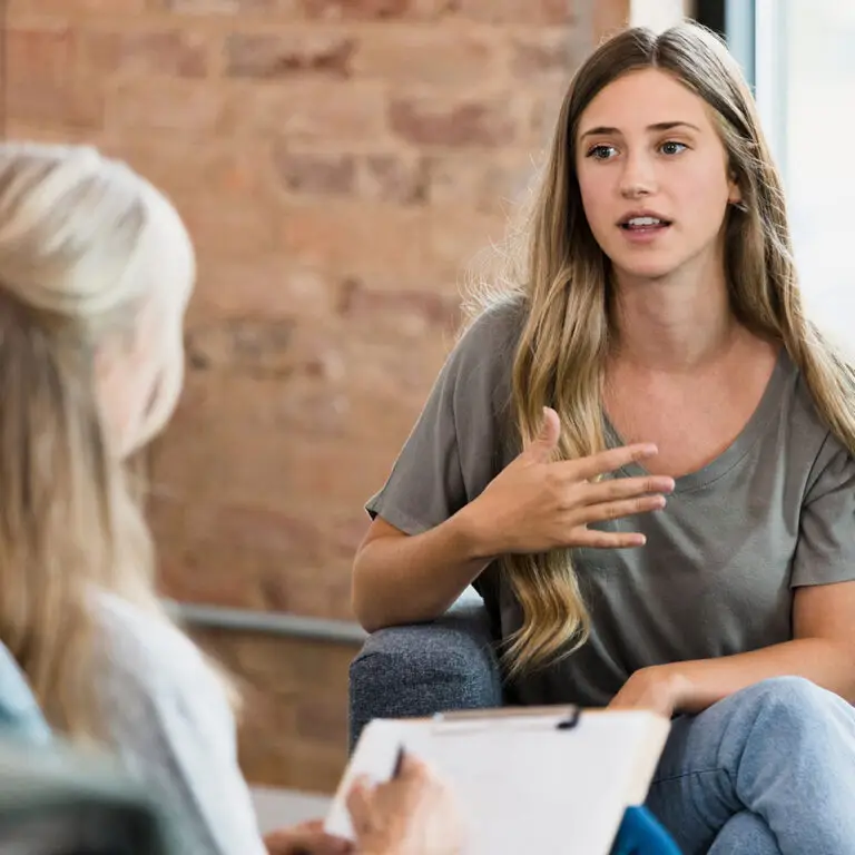 The young adult woman gestures as she talks to the unrecognizable female mature adult counselor.